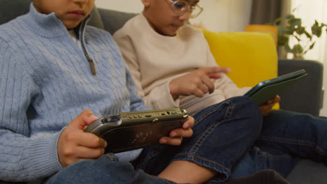 Dos-Niños-Sentados-En-Un-Sofá-En-Casa-Jugando-O-Transmitiendo-En-Una-Tableta-Digital-Y-Un-Dispositivo-De-Juegos-Portátil-4